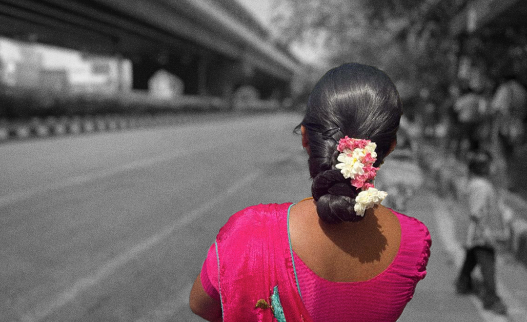 An image of a person in a pink saree and a flowers in her black hair looking away from the camera with the road in the background, used as a representative image for a series on gender-based violence